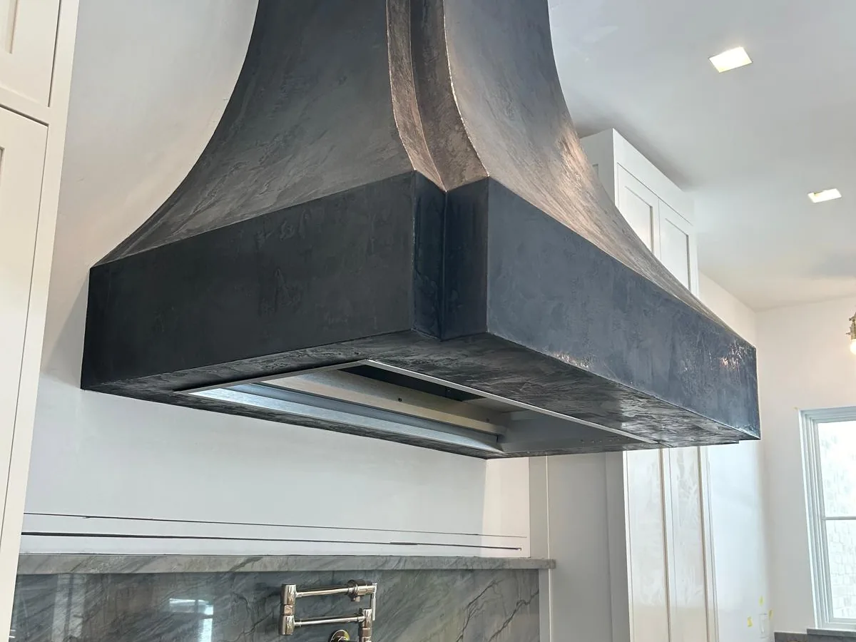 Sculpted dark plaster kitchen range hood over a professional range with a stone backsplash